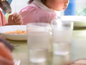 Photo showing a table on which there are two glasses of water and two plates of food and two little girls where one is holding a spoon to eat and the other is blowing on her plate to cool down